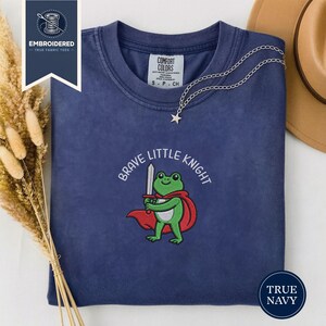 Op de afbeelding: Marineblauw T-shirt met de geborduurde tekst "Brave Little Knight" en een cartoon kikker met een zwaard en rode cape. Een zilveren ketting met een sterhanger ligt op het shirt.