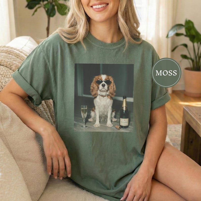 May include: A moss green t-shirt featuring a graphic print of a Cavalier King Charles Spaniel wearing black sunglasses, sitting next to a champagne flute and a bottle of champagne. The text MOSS is printed in a circle on the right side.