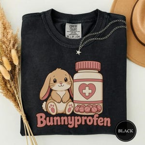 May include: A black short-sleeved t-shirt featuring a graphic of a light brown bunny sitting next to a pill bottle with a heart and cross symbol. The text "Bunnyprofen" is printed in pink below the graphic. The shirt has a Comfort Colors label.