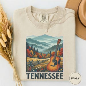 May include: Ivory-colored t-shirt featuring a scenic Tennessee landscape with a guitar. The graphic includes a mountain range, autumn trees, a wooden fence, and the word "Tennessee" in bold letters. The shirt has a crew neck and short sleeves.