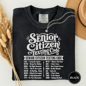 May include: A black crew neck t-shirt featuring white text that reads Senior Citizen Texting Code, followed by a list of humorous acronym definitions such as LOL for Living On Lipitor and BTW for Bring The Walker, printed on the front chest area.