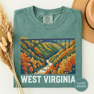 May include: Light green t-shirt featuring a colorful graphic of a bridge over a river in a valley, with autumn foliage. The text "WEST VIRGINIA" is printed in white at the bottom of the graphic. The shirt is made of Comfort Colors material.