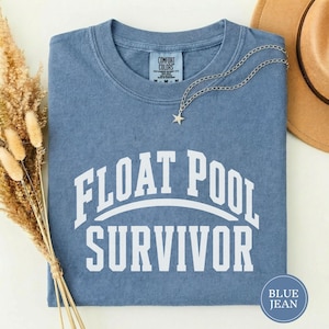 May include: A blue short-sleeved t-shirt with a crew neckline featuring the white text FLOAT POOL SURVIVOR in a collegiate font. The shirt has a Comfort Colors brand label at the inner neck and is accented by a silver-toned star pendant necklace.
