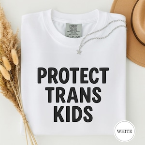 May include: White t-shirt with the words "PROTECT TRANS KIDS" in bold black letters. A silver necklace with a star pendant and a brown hat are visible. The shirt has a Comfort Colors tag.