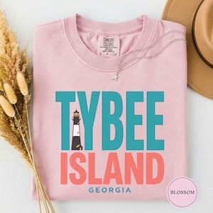 May include: A light pink t-shirt with the words "TYBEE ISLAND GEORGIA" in teal and coral. A black and white lighthouse graphic is centered above the word "ISLAND". A silver necklace with a star pendant is draped across the shirt.