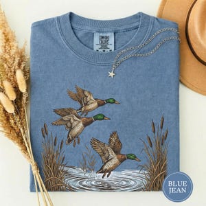 May include: Blue jean colored t-shirt with a graphic of three ducks in flight over water and reeds. The ducks have brown and white feathers and green heads. A silver necklace with a star pendant is on the shirt.