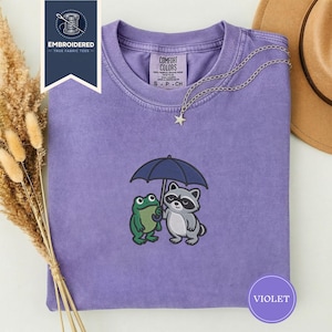 カエルとアライグマが傘を共有している刺繍入りTシャツ、コンフォートカラーズの居心地の良い友情アニマルシャツ、かわいいコテージコアデュオギフトTシャツ