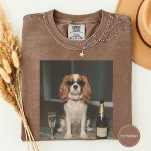 May include: A brown short-sleeved t-shirt with a Comfort Colors label at the neck. The center graphic features a Cavalier King Charles Spaniel wearing black sunglasses, sitting next to a champagne flute and a bottle of champagne with a label.