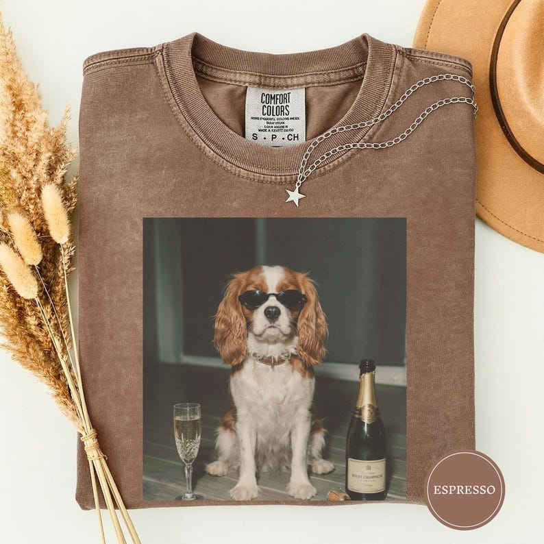 May include: A brown short-sleeved t-shirt with a Comfort Colors label at the neck. The center graphic features a Cavalier King Charles Spaniel wearing black sunglasses, sitting next to a champagne flute and a bottle of champagne with a label.