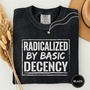 以下が含まれることがあります： 「RADICALIZED BY BASIC DECENCY」という白い文字がプリントされた黒いTシャツ。Tシャツはクルーネックで、首元に小さなタグが付いています。星のペンダントが付いたシルバーのチェーンがTシャツにかけられています。「BLACK」という文字が入った黒い円が下にあります。