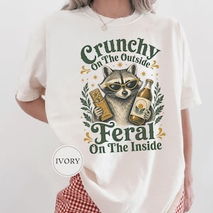 Pode incluir: Uma t-shirt cor de marfim com um gráfico de um guaxinim a usar óculos de sol, segurando uma barra de cereais e uma garrafa de vidro. O texto diz Crunchy On The Outside Feral On The Inside. O design inclui detalhes de folhas verdes e douradas.