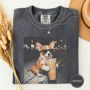 May include: Dark gray t-shirt featuring a graphic of a corgi wearing sunglasses and drinking an iced coffee. The shirt has a crew neck and short sleeves. A silver necklace with a star pendant is draped on the shirt. The tag reads "COMFORT COLORS".