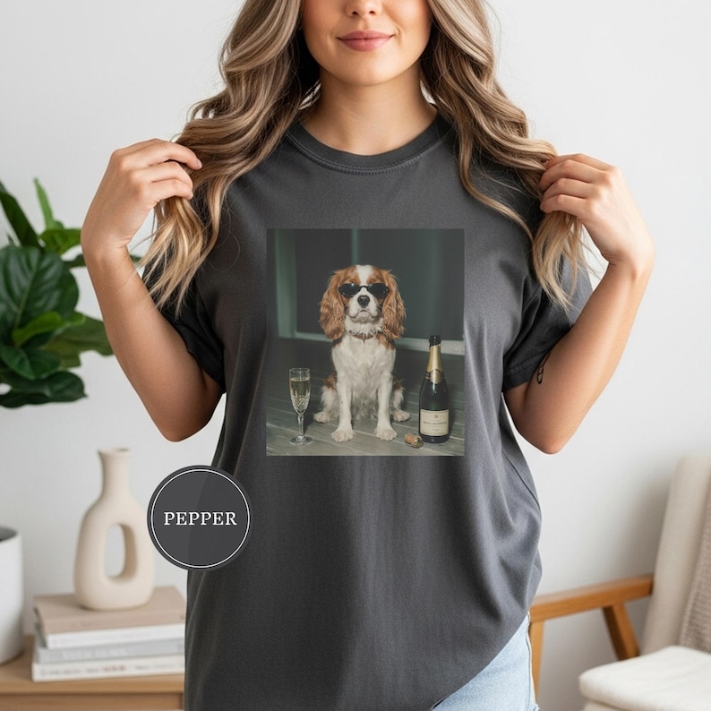 May include: A charcoal gray t-shirt featuring a graphic print of a Cavalier King Charles Spaniel wearing black sunglasses, sitting next to a champagne flute and a bottle of champagne. A circular label with the text PEPPER is on the lower left.