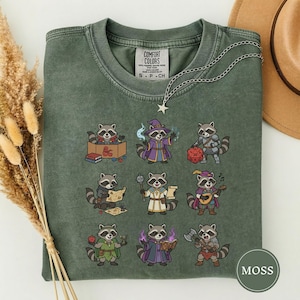 May include: Moss green Comfort Colors t-shirt featuring a collection of cartoon raccoons in various fantasy roles. The shirt includes a silver necklace with a star pendant. The text "COMFORT COLORS" is visible on the shirt.