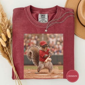 Op de afbeelding: Karmizijnrood T-shirt met een eekhoorn in een honkbaluniform. De eekhoorn draagt een rode pet en zonnebril en houdt een knuppel vast. Het shirt heeft een Comfort Colors-label en een zilveren ketting met een sterhanger.