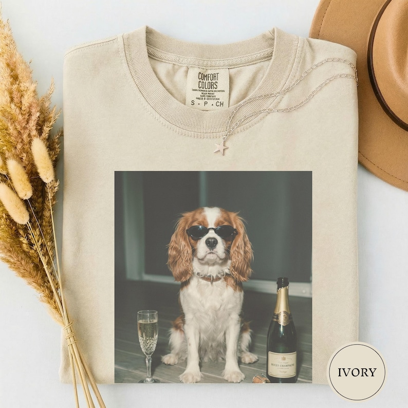May include: An ivory crew neck t-shirt featuring a graphic print of a brown and white spaniel dog wearing black sunglasses, sitting next to a champagne flute and a bottle of champagne. The shirt has a Comfort Colors brand label at the neck.