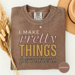 May include: Espresso-colored t-shirt with the text "I MAKE PRETTY THINGS permanent" in pink, gold, and green. A silver necklace with a star pendant rests on the shirt. The shirt is folded, and a hat is visible in the background.