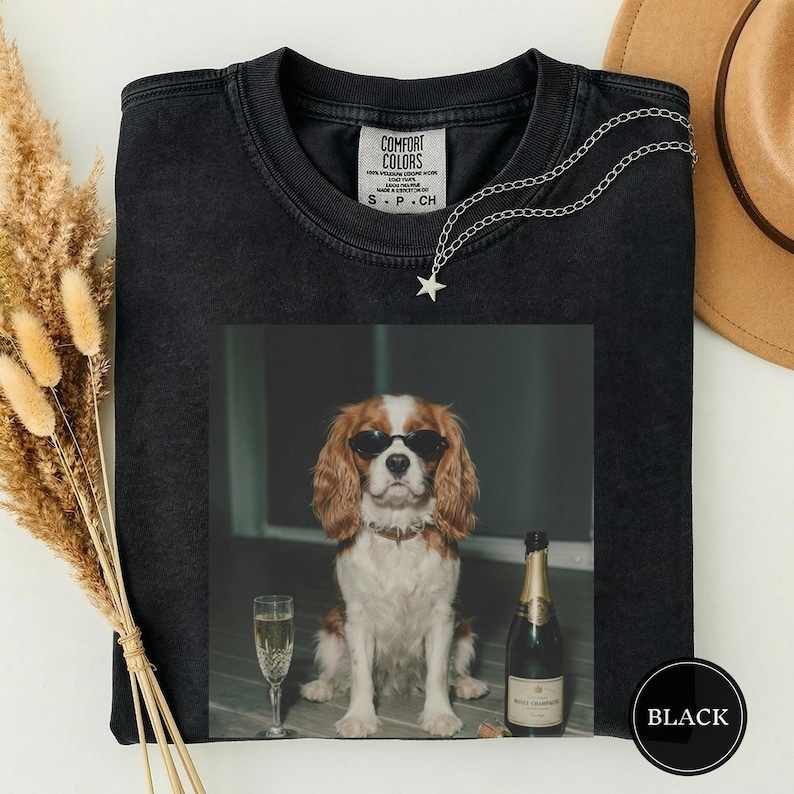May include: A black crew neck t-shirt featuring a graphic print of a brown and white Cavalier King Charles Spaniel wearing black sunglasses, sitting next to a champagne flute and a bottle of champagne. The shirt has a Comfort Colors brand label.