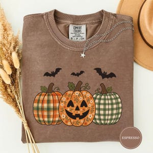 May include: A brown crewneck t-shirt featuring a graphic of three pumpkins and three black bats. The left pumpkin has an orange plaid pattern, the center pumpkin is a jack-o'-lantern with a floral print, and the right pumpkin has a green plaid pattern.