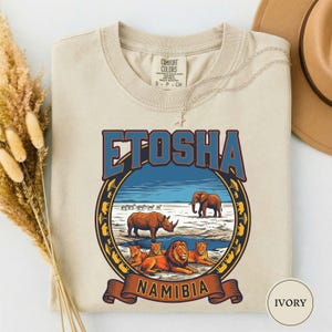 Op de afbeelding: Ivoor kleurig t-shirt met een afbeelding van Etosha National Park in Namibië. Het ontwerp bevat het woord "ETOSHA" in blauw, een neushoorn, een olifant, leeuwen en het woord "NAMIBIA" op een banner.