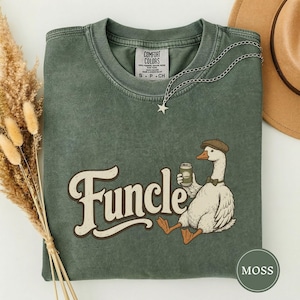 May include: A moss green crewneck t-shirt featuring a graphic of a white goose wearing a brown cap and bow tie while holding a beverage can. The text Funcle is printed in a large cream-colored script font. The shirt has a Comfort Colors label.