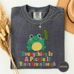 Op de afbeelding: Een antracietgrijs t-shirt met korte mouwen en een afbeelding van een groene kikker die een groene augurk vasthoudt. De tekst Everything Is A Pickle If You're Brave Enough staat in kleurrijke letters. Het Comfort Colors label zit in de nek.