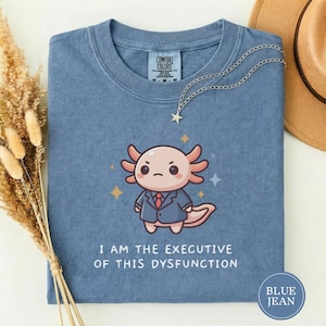 Puede incluir: Camiseta azul vaquero con un ajolote de dibujos animados con traje y corbata. El texto "I AM THE EXECUTIVE OF THIS DYSFUNCTION" está impreso debajo. Un collar plateado con un colgante de estrella está sobre la camiseta.