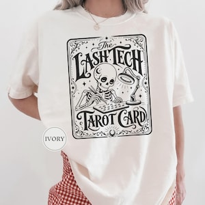 May include: An ivory t-shirt featuring a black graphic print of a skeleton working with lash tools under a lamp. The design is framed like a tarot card with the text The LASH TECH TAROT CARD. A small circular label on the shirt reads IVORY.