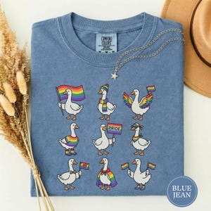 May include: Blue jean coloured t-shirt with a crew neck. The shirt features a design of cartoon geese with rainbow flags, scarves, and accessories. The shirt has the words "Comfort Colors" on the tag.
