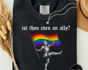 May include: Black t-shirt with the text "ist thou even an ally?" in white gothic font. The shirt features a graphic of a knight holding a rainbow flag. A silver chain with a star pendant is draped across the shirt.