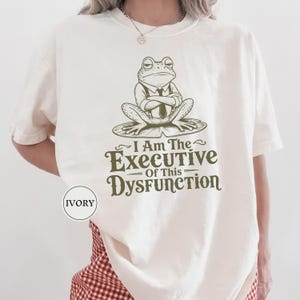 May include: An ivory t-shirt featuring a dark green graphic of a frog wearing a shirt, tie, and suspenders while sitting on a lily pad. Below the frog, the text reads, I Am The EXECUTIVE Of This DYSFUNCTION. A small circular label says IVORY.