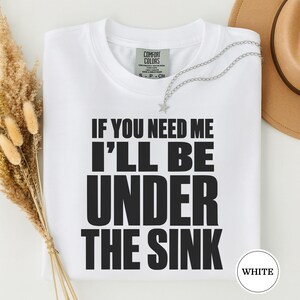 Könnte beinhalten: Ein weißes kurzärmeliges T-Shirt mit dem schwarzen Text IF YOU NEED ME I'LL BE UNDER THE SINK auf der Vorderseite. Das T-Shirt hat ein Comfort Colors Markenetikett am inneren Halsausschnitt und wird mit einer silbernen sternförmigen Halskette gezeigt.