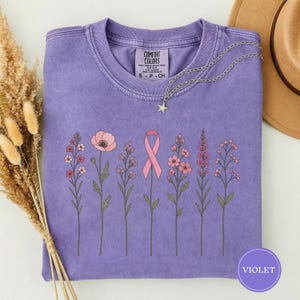 May include: A violet short-sleeved t-shirt featuring a graphic print of six botanical stems and a pink awareness ribbon in the center. The shirt has a crew neckline with a Comfort Colors label. A silver star-shaped pendant necklace rests on top.