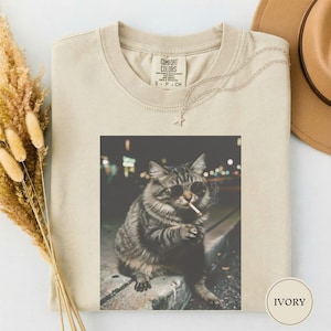 May include: Ivory-colored Comfort Colors t-shirt with a graphic of a cat wearing sunglasses and smoking a cigarette. The shirt has a crew neck and short sleeves. A gold-colored necklace with a star pendant is draped on the shirt.