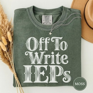 Könnte beinhalten: Ein moosgrünes Kurzarm-T-Shirt mit Rundhalsausschnitt und einem weißen Grafikdruck, der in dekorativer Schrift "Off To Write IEPs" lautet. Das T-Shirt hat ein Comfort Colors Markenetikett am Innenhals und eine silberne Stern-Halskette.
