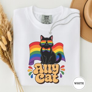 May include: White t-shirt with a black cat wearing rainbow sunglasses and a bow tie, set against a rainbow flag. The text "Any Cat" is below the cat. A silver necklace with a star pendant is on the shirt.