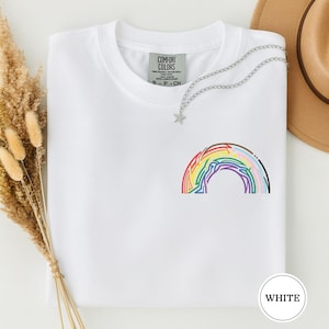 Puede incluir: Una camiseta blanca con un pequeño diseño de arcoíris bordado en varios colores. La camiseta tiene cuello redondo y una etiqueta que dice "COMFORT COLORS". Un collar plateado con un colgante de estrella está drapeado sobre la camiseta.