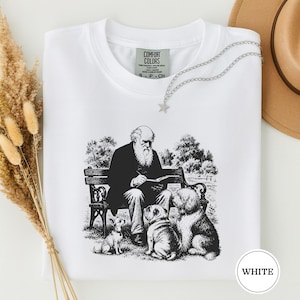 May include: A white t-shirt featuring a black ink illustration of a bearded man sitting on a park bench writing in a notebook, surrounded by three dogs. The shirt has a Comfort Colors label at the neck and a silver star-shaped necklace.