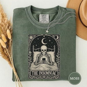 May include: A moss green crewneck t-shirt featuring a graphic print of a skeleton lying in bed using a smartphone under a crescent moon and stars. The text THE INSOMNIAC is printed below the illustration. The shirt has a Comfort Colors label.