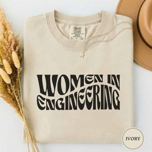 以下が含まれることがあります： アイボリー色のTシャツ。黒い波状のフォントで「WOMEN IN ENGINEERING」の文字がプリントされています。クルーネックと半袖です。星のペンダントが付いたシルバーのネックレスと茶色の帽子も写っています。