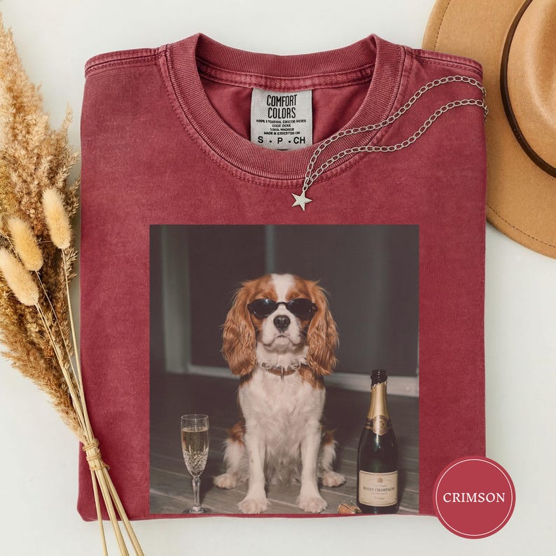 May include: A crimson crewneck t-shirt featuring a graphic print of a brown and white spaniel dog wearing black sunglasses, sitting next to a champagne flute and a bottle of champagne. The shirt has a Comfort Colors label at the inner neckline.