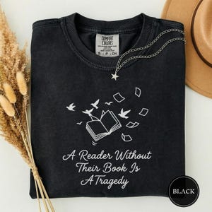 Op de afbeelding: Zwart T-shirt met witte afbeelding van een open boek, vogels en zwevende pagina's. De tekst "A Reader Without Their Book Is A Tragedy" staat eronder gedrukt. Een zilveren ketting met een sterhanger ligt op het T-shirt.