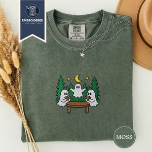 May include: Moss green t-shirt with an embroidered design of three ghosts around a table under a crescent moon and stars. The shirt has a Comfort Colors tag and a silver chain with a star pendant. The shirt is folded with a brown hat visible.