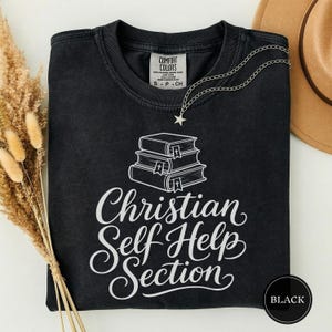 Puede incluir: Una camiseta negra de cuello redondo con un gráfico blanco de una pila de tres libros con símbolos de cruces en los lomos. Debajo de los libros, el texto "Christian Self Help Section" aparece en letra cursiva. Etiqueta Comfort Colors.