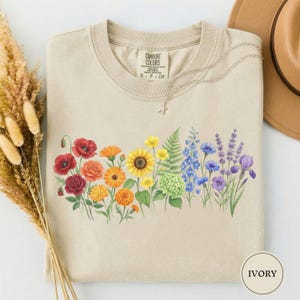 May include: Ivory-coloured t-shirt with a watercolour floral design. The design features a rainbow of flowers, including red poppies, orange marigolds, yellow sunflowers, and blue delphiniums. The shirt has a crew neck and short sleeves.