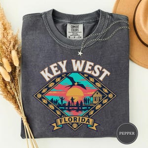 Könnte beinhalten: Dunkelgraues T-Shirt mit einem farbenfrohen Key West, Florida-Motiv. Das Design zeigt eine Sonnenuntergangsszene mit einem Vogel, Menschen auf einem Pier und einem Haus. Das Shirt hat einen Rundhalsausschnitt und ein Etikett mit der Aufschrift "Comfort Colors". Eine silberne Halskette mit Sternanhänger liegt auf dem Shirt.