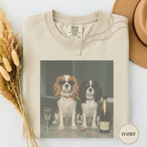Op de afbeelding: Ivoor Comfort Colors T-shirt met een afbeelding van twee Cavalier King Charles Spaniels met zonnebrillen, een champagnefles en een glas. Het shirt heeft een ronde hals en korte mouwen. Een zilveren ketting en een bruine hoed zijn ook zichtbaar.