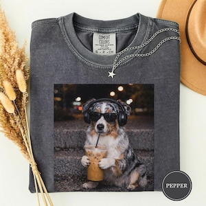 May include: Charcoal grey Comfort Colors t-shirt with a graphic of a dog wearing sunglasses and headphones, holding an iced coffee. A silver chain with a star pendant and a brown hat are also visible.