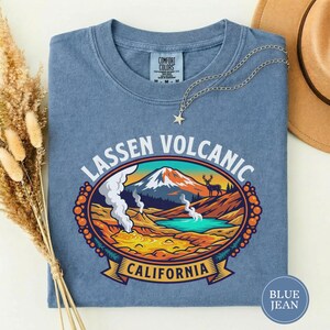 May include: Blue jean-coloured t-shirt featuring a graphic of Lassen Volcanic National Park. The design shows a mountain, lake, and deer, with the text "Lassen Volcanic California". A silver necklace with a star pendant is on the shirt.