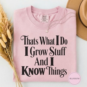 May include: A light pink short-sleeved t-shirt featuring the black printed text "Thats What I Do I Grow Stuff And I Know Things". The shirt has a crew neckline and a Comfort Colors brand label on the inside collar. A silver star necklace is visible.